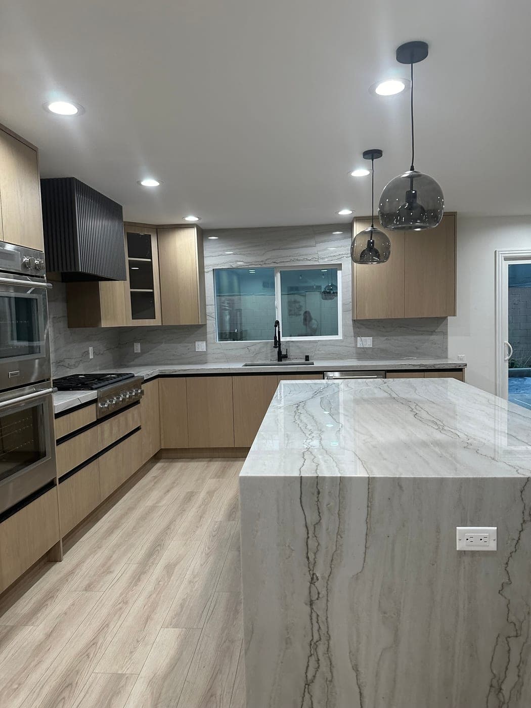 Quartzite waterfall island under smoked glass pendants, with the oak sink wall behind