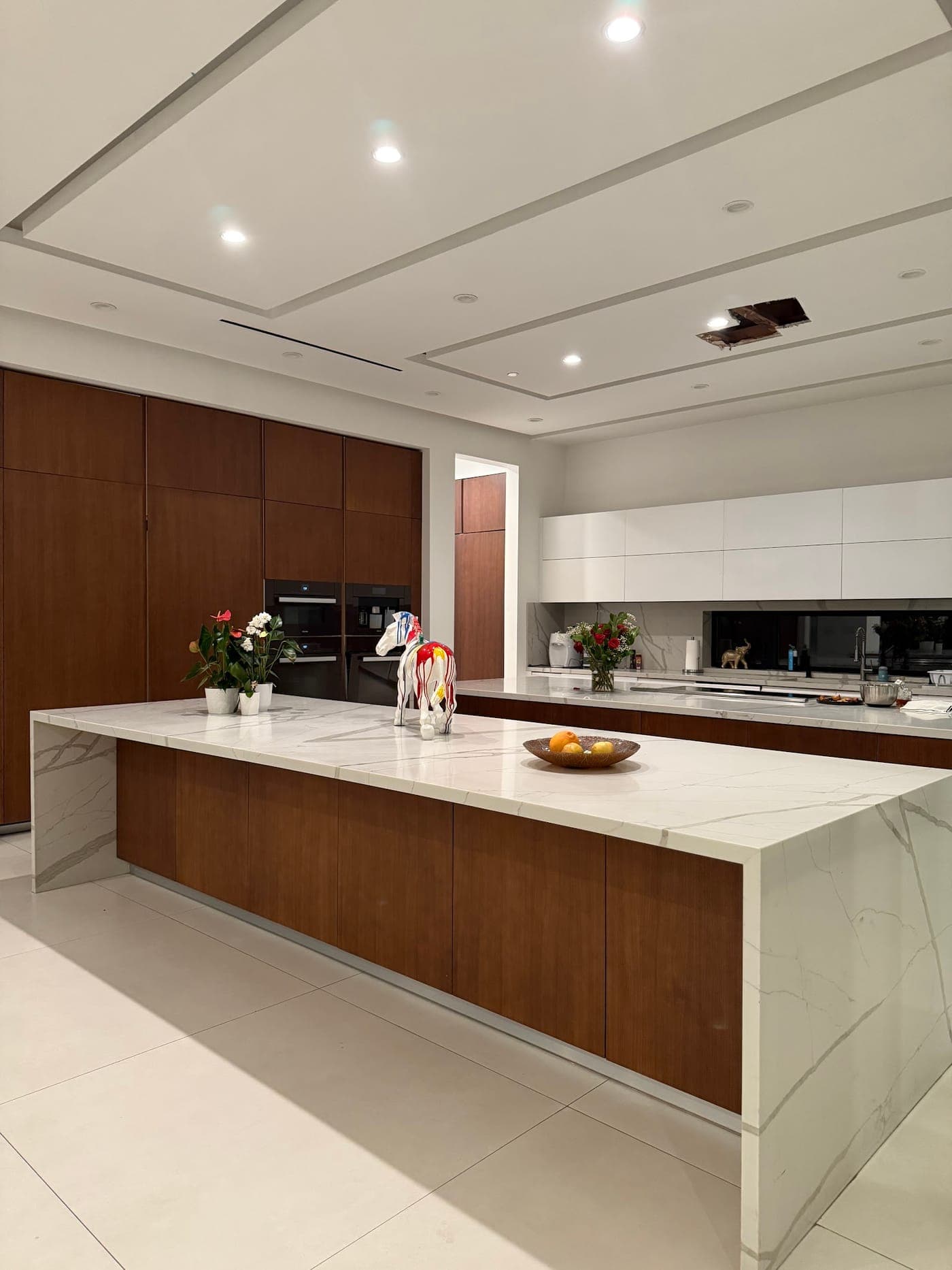 Long island with a white veined waterfall countertop over walnut-tone flat-panel cabinets, with tall wood cabinets and built-in ovens behind