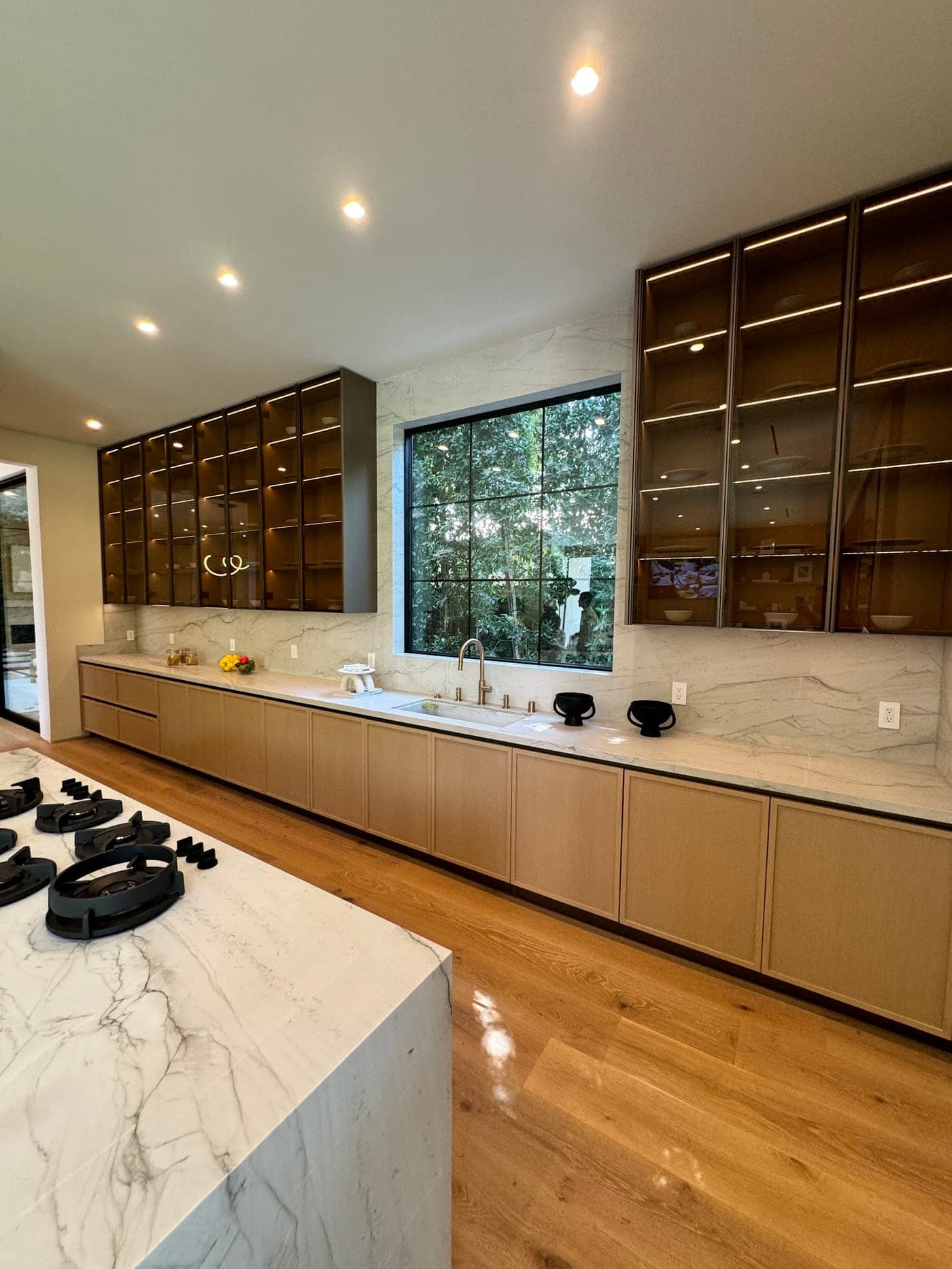 Long run of light oak flat-panel cabinets under a veined backsplash, with lit glass-front cabinets above and the cooktop island in front