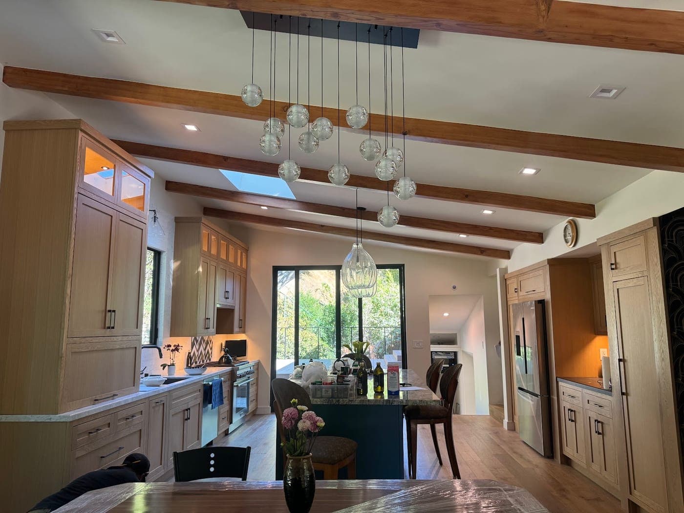 Open kitchen with oak cabinetry under exposed wood beams and a cluster of glass pendant lights
