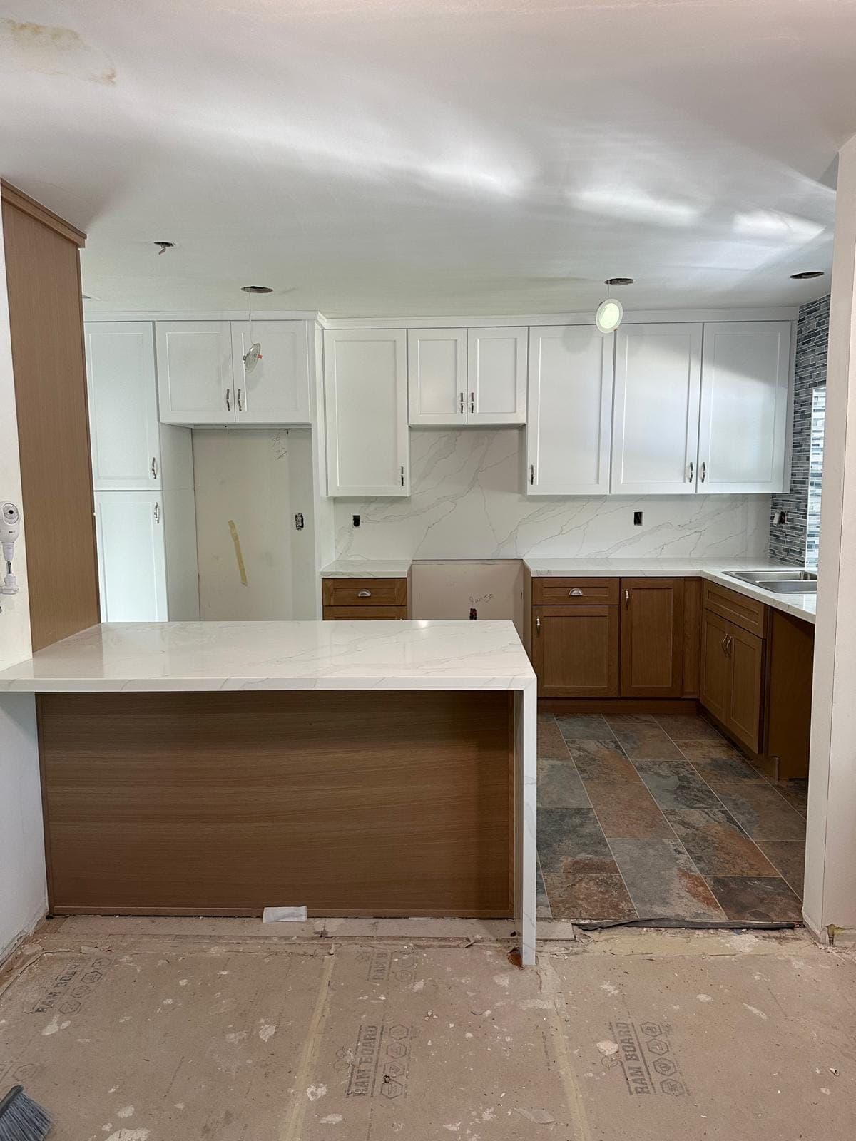 White upper cabinets, wood-tone base cabinets and a peninsula with a waterfall countertop, near the end of the work
