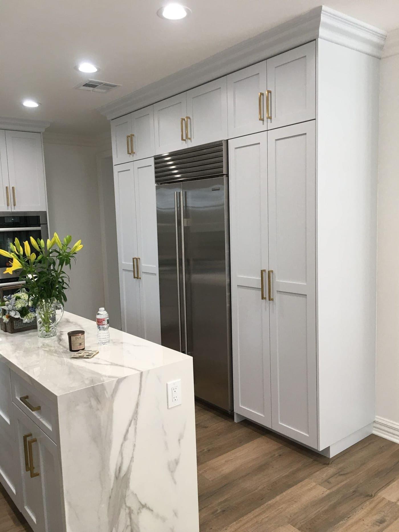 White Shaker cabinets with brass pulls around a built-in stainless refrigerator, and an island with a white veined waterfall edge